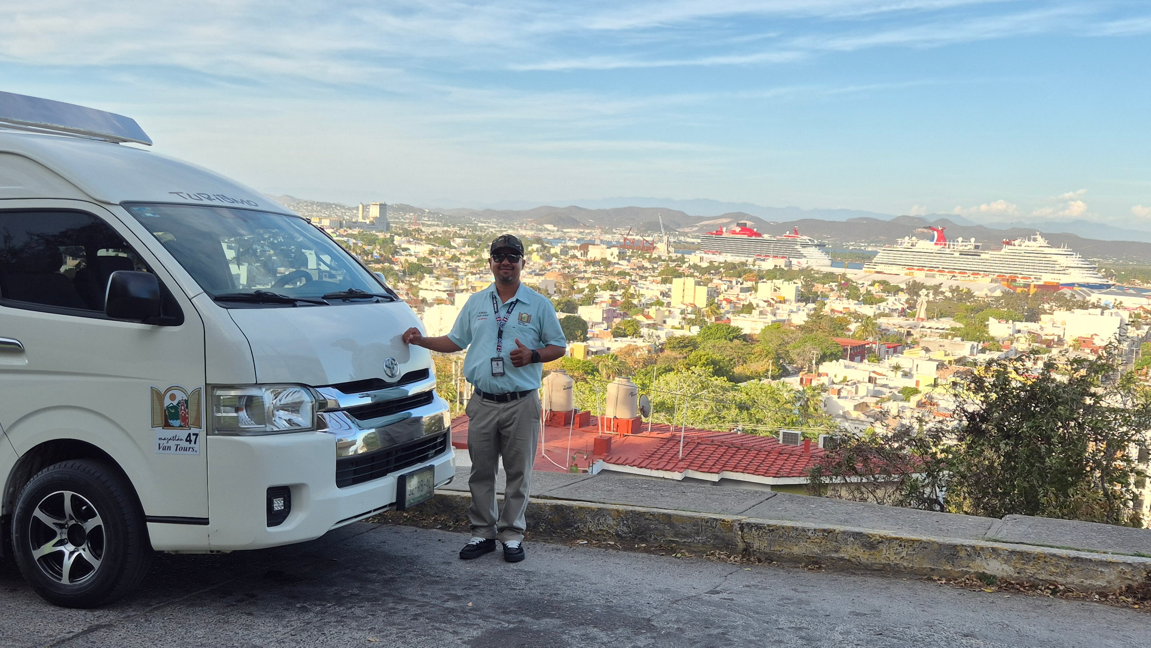 Guía de turistas certificado SECTUR junto a su van turística blanca en un mirador con la bahía y cruceros de Mazatlán al fondo